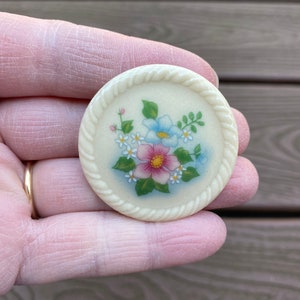 May include: A white ceramic brooch with a floral design. The flowers are pink, blue, and white with green leaves. The brooch has a rope-like border.