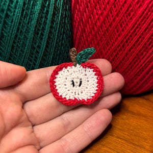 May include: A small, circular crochet apple slice applique featuring a white center, a red outer border, a brown stem, and a single green leaf. The design includes two small black embroidered stitches in the center to represent apple seeds.