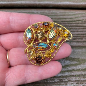 May include: A gold-tone leaf-shaped brooch featuring a textured metal frame set with various round and marquise-cut rhinestones in shades of amber, brown, and iridescent multi-colored hues, arranged in a cluster across the openwork metal surface.