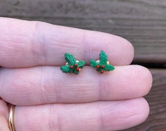 May include: A pair of small, green holly leaf earrings with red berry accents and gold-colored posts. The earrings are designed to resemble a sprig of holly, a common Christmas decoration.