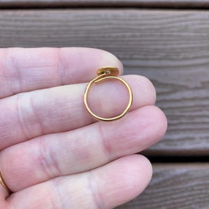 Op de afbeelding: Een goudkleurige ring met een klein, cirkelvormig detail. De ring wordt in een hand gehouden, wat het delicate ontwerp en de gepolijste afwerking benadrukt. De ring heeft een dunne, eenvoudige band met een klein decoratief element.