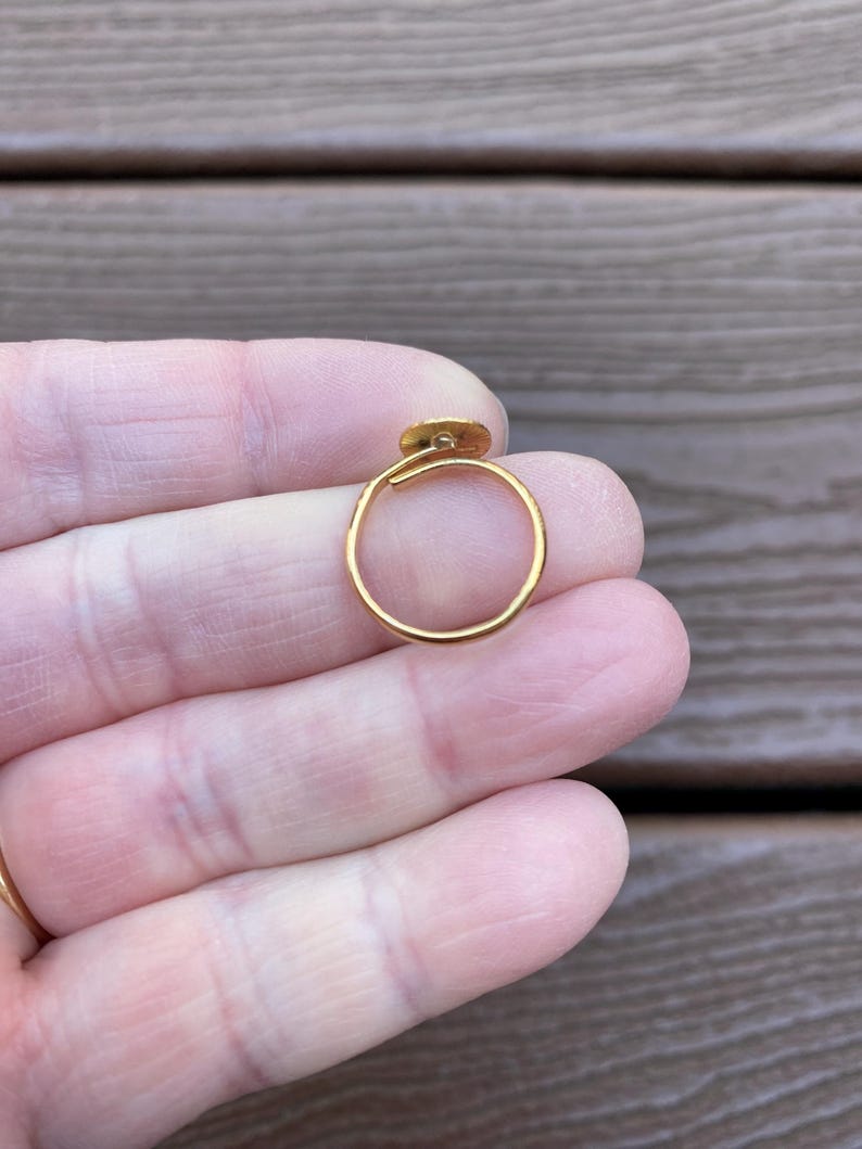 Op de afbeelding: Een goudkleurige ring met een klein, cirkelvormig detail. De ring wordt in een hand gehouden, wat het delicate ontwerp en de gepolijste afwerking benadrukt. De ring heeft een dunne, eenvoudige band met een klein decoratief element.