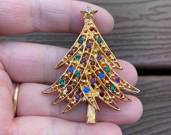 May include: Gold-toned Christmas tree brooch with colorful gemstone accents. The brooch features a star at the top and is adorned with red, green, blue, and purple gems. A festive accessory for the holiday season.