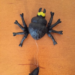 May include: A black plastic spider toy with eight legs and a yellow head. A string is attached to the spider, leading to a small black bell. The toy is likely for Halloween or a similar holiday.