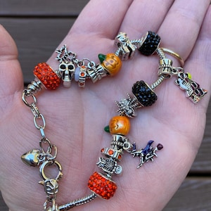 May include: A silver charm bracelet with Halloween-themed charms, including a skull, pumpkins, a witch, and a haunted house. The bracelet is adorned with red and orange crystal beads.