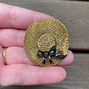 Könnte beinhalten: Eine goldfarbene Brosche in Form eines Strohhutes mit einer schwarzen Schleife und grünen und goldenen Akzenten.