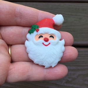 May include: A Christmas-themed Santa Claus face ornament. The ornament has a red hat with a white bobble, a white beard, rosy cheeks, and a smiling red mouth. A sprig of holly with red berries decorates the hat.