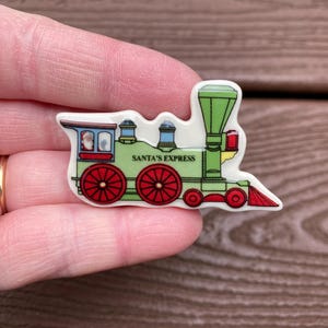 Puede incluir: Un pin colorido con forma de tren navideño. El pin presenta una locomotora verde con ruedas y detalles rojos, y el texto "SANTA'S EXPRESS". El tren tiene un frente rojo y blanco y una pequeña ventana con una figura de Papá Noel.