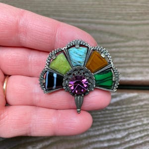 May include: A fan-shaped brooch featuring a central round purple faceted stone surrounded by five trapezoidal inlay pieces in black, lime green, light blue, amber, and dark green, all set within a textured silver-tone metal frame.
