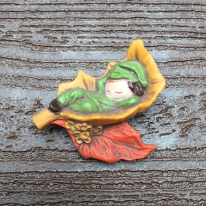 May include: A ceramic figurine of a sleeping child nestled in a green, orange, and yellow leaf. The child is wearing a green hat and has brown hair.