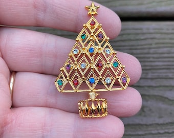 May include: A gold-tone Christmas tree brooch with a star on top and a base. The tree is decorated with colorful rhinestones in red, blue, green, and clear.