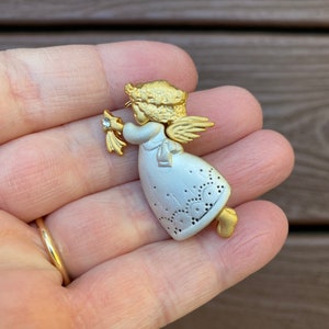 May include: Gold and silver tone angel brooch with a white dress and gold wings. The angel is holding a small flower with a rhinestone in the center.