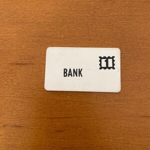 Peut inclure: Une carte rectangulaire blanche avec le mot "BANK" imprimé en noir, ainsi qu'un graphique de timbre noir avec le chiffre "1". La carte est posée sur une surface en bois brun.