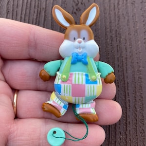Può includere: Un colorato giocattolo a forma di coniglietto pasquale con orecchie e zampe marroni, un papillon blu e una camicia verde. Il coniglietto indossa pantaloni patchwork con quadrati rosa, blu e gialli. Un filo verde è attaccato a un bottone azzurro.