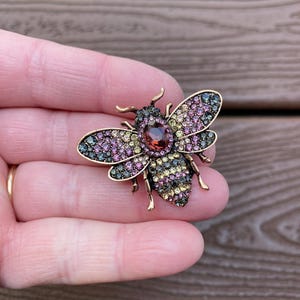 May include: A gold-toned bee brooch with a central oval-shaped red gemstone and wings adorned with small, colorful gemstones in shades of pink, green, and yellow. The brooch is held against a wooden background.