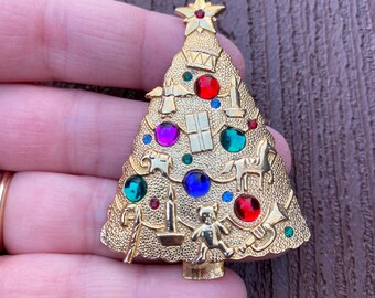 May include: Gold-toned Christmas tree brooch with colorful jewel accents. The brooch features a star on top and is adorned with red, green, blue, and purple jewels. Decorative elements include a teddy bear, candle, and gift.