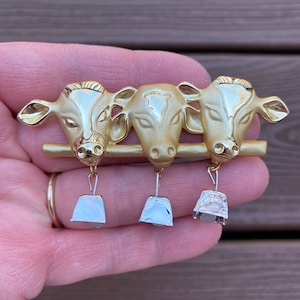 Puede incluir: Broche de metal dorado con tres cabezas de vaca en fila. Cada cabeza de vaca tiene una campana colgando debajo. El broche se sostiene en una mano, mostrando los intrincados detalles de las cabezas y campanas.