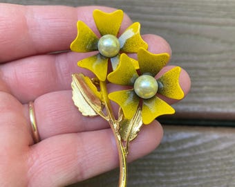 Peut inclure: Une broche en métal doré représentant deux fleurs émaillées jaunes avec des ombres foncées au centre. Chaque fleur possède une perle artificielle blanche en son milieu. La broche comprend une tige dorée avec deux feuilles texturées.