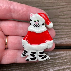 May include: A ceramic brooch shaped like a white cat with black stripes, wearing a red Santa hat and a red coat with white trim. The cat has green eyes and a pink nose, with a glossy finish and visible fine crackle patterns in the white glaze.