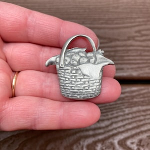 May include: A silver-coloured metal brooch shaped like a woven basket filled with rounded items. The basket features a textured, cross-hatched pattern on the exterior, a single arched handle, and a smooth, draped fabric element extending over the side.