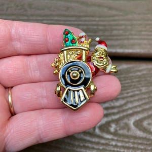 May include: A gold-tone metal brooch shaped like a steam locomotive engine. The train features black enamel detailing on the front, a green Christmas tree with red accents on top, and a figure of Santa Claus in a red suit attached to the side.