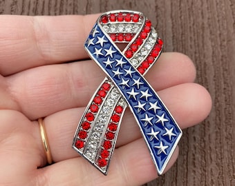 May include: A ribbon-shaped brooch featuring a metallic finish with a patriotic design. One side displays a deep blue enamel background with silver-toned stars, while the other side is adorned with alternating rows of red and clear crystals.