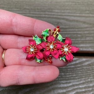 Könnte beinhalten: Eine Brosche aus goldfarbenem Metall mit drei leuchtend roten Weihnachtssternblüten mit strukturierten Blütenblättern, die jeweils in der Mitte mit einem klaren Strassstein besetzt sind. Das Design umfasst grüne Emaille-Stechpalmenblätter.