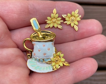May include: A gold-tone teacup and saucer brooch with a white base and pink and blue polka dots. A gold-tone spoon is in the teacup. A yellow flower with gold leaves is on the side of the teacup. Two matching yellow flower earrings with gold leaves are included.