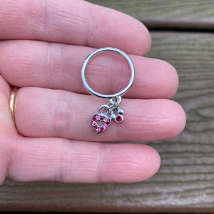 Puede incluir: Un anillo de plata con un dije de coraz&oacute;n rosa y dos peque&ntilde;os acentos de gemas rosas.