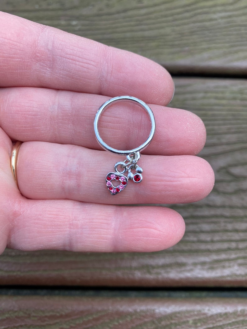 Puede incluir: Un anillo de plata con un dije de coraz&oacute;n rosa y dos peque&ntilde;os acentos de gemas rosas.