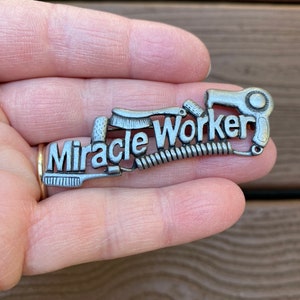 Puede incluir: Encanto plateado con las palabras "Miracle Worker" y varias herramientas para peinar el cabello, incluyendo un secador de pelo, un peine y una plancha para rizar.