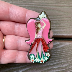 May include: A brooch featuring a stylized figure with pink hair and wings, wearing a red glittery skirt over a green base. The figure holds a small white bird above its head, and the bottom of the piece is decorated with several small white flowers.