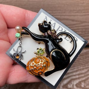 May include: Black cat brooch with a pumpkin and spider design. The cat is black with gold accents, holding a pearl and a spider charm. The pumpkin is orange with green leaves. The brooch is on a white and gray card.