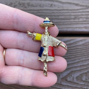 Peut inclure: Broche épouvantail en métal doré avec un chapeau de paille et des patchs colorés. L'épouvantail a les bras tendus et un design détaillé, avec des accents jaunes, bleus et rouges. Un accessoire charmant.
