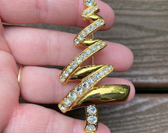 May include: Gold-tone Christmas tree brooch with clear rhinestone accents. The brooch features a stylized, spiral design with a series of sparkling stones. The tree has a rounded base and a single rhinestone at the top.