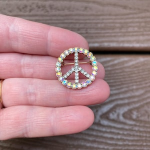 以下が含まれることがあります： イリデッセントラインストーンが付いた、金色の平和のシンボルブローチ。平和のシンボルはカラフルな石のリングで縁取られ、内側の線はクリアラインストーンで飾られています。ヴィンテージスタイルのアクセサリー。