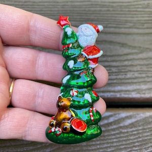 May include: A metal brooch shaped like a green Christmas tree with white snow accents. A Santa Claus figure in a red suit is at the top placing a red star, while a brown teddy bear sits at the base with red and white candy cane decorations.