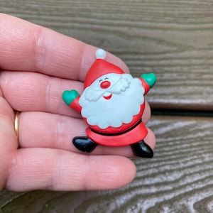 Puede incluir: Un broche de Papá Noel con gorro rojo y pompón blanco, barba blanca, nariz roja, manoplas verdes y botas negras. La figura se muestra en una pose alegre con los brazos extendidos, elaborada con superficies lisas y de colores sólidos.