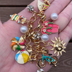 Könnte beinhalten: Ein goldfarbenes Charm-Armband mit sommerlichen Anhängern. Zu den Anhängern gehören ein Segelboot, Flip-Flops, eine Sonne, ein Fahrrad, ein Strandball und ein Windrad. Akzentuiert mit Kunstperlen und kristallartigen Verzierungen.