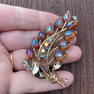 May include: A vintage gold-tone brooch in the shape of a floral bouquet. The brooch features iridescent, teardrop-shaped stones in shades of orange, blue, and purple. The brooch has a detailed design with a bow and leaves.