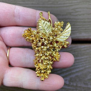 Könnte beinhalten: Eine goldfarbene Brosche in Form eines Blumenbüschels mit Blättern. Die Blüten sind klein und zart, die Blätter groß und strukturiert.