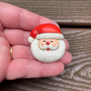 May include: A Santa Claus brooch featuring a bright red hat with a white textured brim, a peach-coloured face, a white textured beard and moustache, and a small red nose. The character has closed eyes and a smiling mouth with a small red accent.