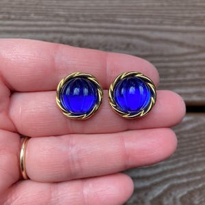 May include: A pair of vintage earrings featuring a gold-tone twisted rope design surrounding a vibrant, domed, cobalt blue glass center. The earrings are round and have a classic, elegant style.