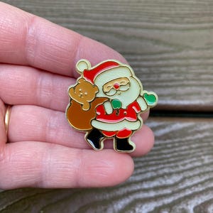 May include: A gold-toned metal enamel pin featuring Santa Claus wearing a red suit with white trim, black boots, and green mittens. Santa is carrying a brown sack over one shoulder with a small brown teddy bear peeking out from the top of the bag.