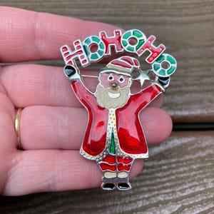 May include: A silver-tone metal brooch featuring a Santa Claus figure with red, white, and green enamel details. Santa is depicted with arms raised, holding a banner above the head that reads HOHOHO in red and green lettering with a small star.