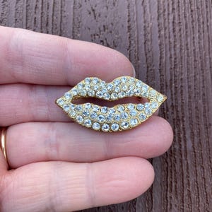 以下が含まれることがあります： クリアなラインストーンで覆われた唇の形をしたゴールドトーンのブローチ