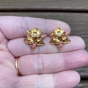 Könnte beinhalten: Ein Paar goldfarbene blumenförmige Ohrringe mit metallischem Finish, zarten rosa Emaille-Akzenten auf den Blütenblättern und einer kleinen weißen Kunstperle in der Mitte jeder Blüte, gestaltet als Ohrstecker mit einem floralen Design.