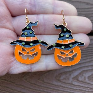 May include: A pair of Halloween-themed earrings. Each earring features a smiling orange pumpkin wearing a black witch's hat with green accents. The earrings have gold-colored hooks.