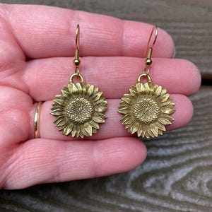 Könnte beinhalten: Ein Paar goldfarbene Ohrhänger mit einem detaillierten Sonnenblumenmotiv. Jeder Ohrring besteht aus einem runden Sonnenblumenanhänger mit strukturierten Blütenblättern und einem zentralen Blütenkopf, der an einem einfachen Metallhaken hängt.