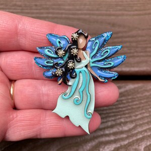 May include: A fairy-themed brooch with a light blue dress and matching wings. The fairy has black hair and is adorned with small white flower accents. The wings have a glittery blue outline. The brooch is a whimsical accessory.